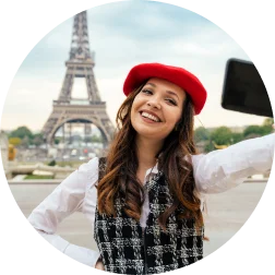Une femme souriante, coiffée d'un béret rouge et vêtue d'une veste noire, prend un selfie avec la tour Eiffel en arrière-plan. La scène est lumineuse et joyeuse, évoquant le charme parisien.
