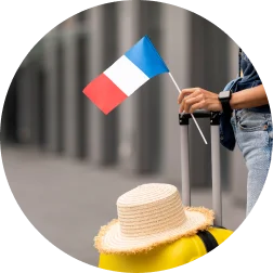 Une personne vêtue d'un jean tient un petit drapeau français, debout à côté d'une valise jaune sur laquelle est posé un chapeau de paille, suggérant un voyage. L'arrière-plan est urbain et flou.