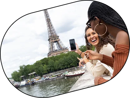 Deux femmes souriantes profitent de la vue sur la Tour Eiffel depuis les berges de la Seine à Paris. L'une montre son téléphone à l'autre. L'ambiance est joyeuse et détendue.